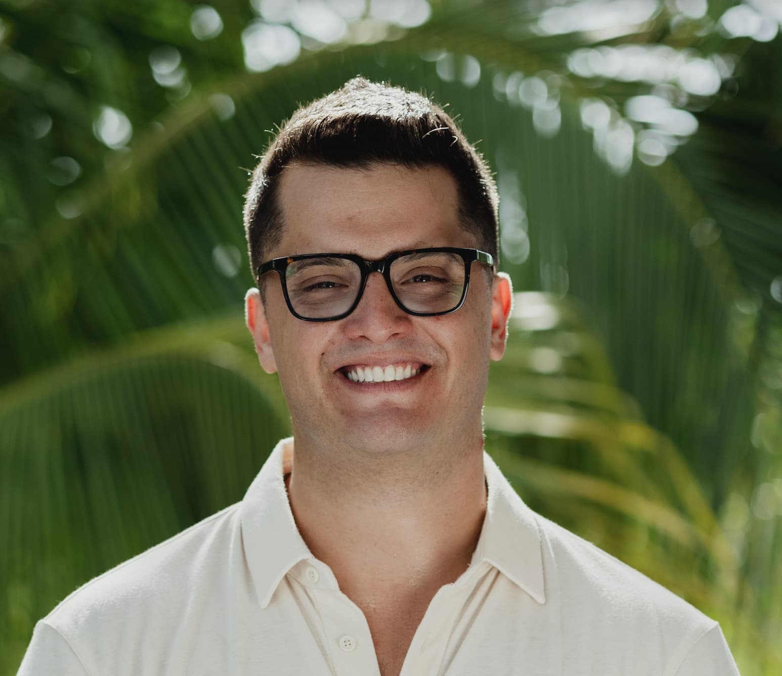 Nik Vassev smiling outdoors in a cream polo and glasses, with tropical palms behind him.